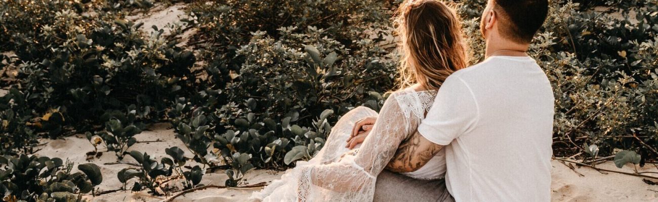 A vertical shot of a man and a woman sitting and hugging in a field while looking at the sunset